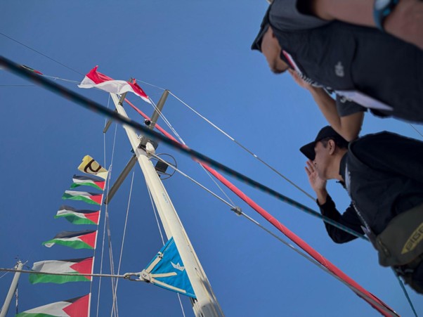 Two people on a sailboat beside a tall mast with multicolored signal flags and rigging against a clear blue sky.