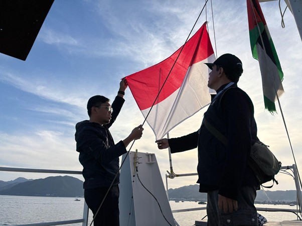 Two men on a boat hoist a red-and-white flag (Indonesia) under a blue, cloudy sky with mountains in the distance.