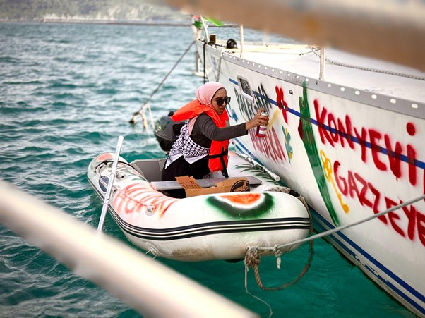 Person in a red life jacket and hijab spray-paints colorful graffiti on a large boat while seated in an inflatable dinghy beside it.