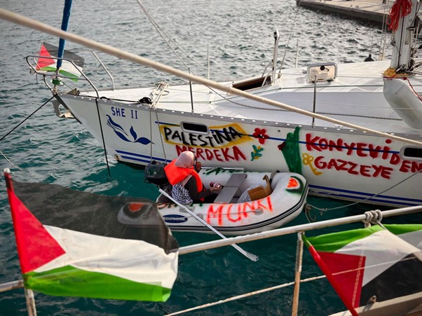 Small dinghy with an orange life jacket beside a graffiti-painted sailboat reading 'Palestina' and 'Gaza' in a harbor, with Palestinian flag bunting in the foreground.