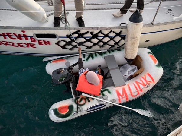 Small dinghy with an outboard motor tied to a sailboat; graffiti reads 'MUNKY' and other red letters on the hull; a person wearing an orange life jacket sits in the dinghy surrounded by gear over green water.
