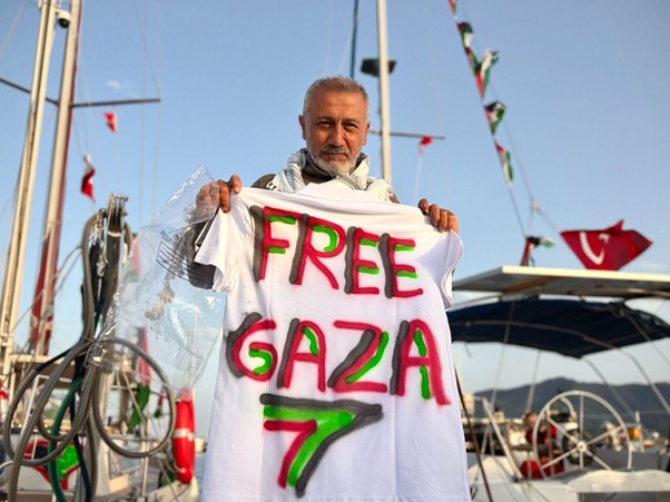 Man at a marina holding a white T‑shirt with bold red and green letters reading 'FREE GAZA' in front of boats and sailing equipment