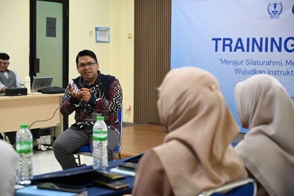 Instructor speaks into a microphone during a training session; attendees in hijabs sit at desks while a blue TRAINING banner appears in the background.