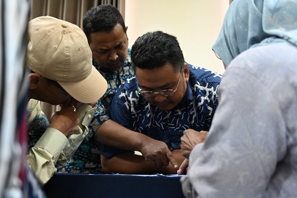 Group of men leaning over a table, closely examining a small object together in a crowded setting.