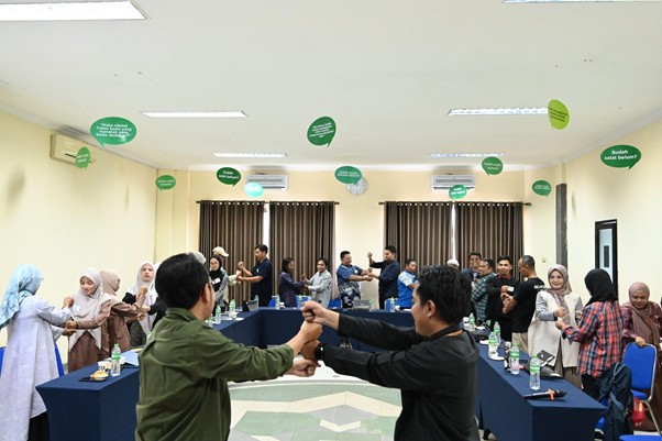 Group of people in a meeting room participate in a team-building activity, standing in a circle and linking arms, with green speech-bbubble decorations overhead.