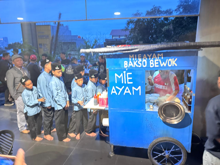 vespa60sjabodetabekdanscomadiindonesia3 dompet dhuafa Line of barefoot students in blue jackets and black hats queue at a blue street food cart labeled 'Mie Ayam' and 'Bakso Be Wok' with a cook inside preparing food.