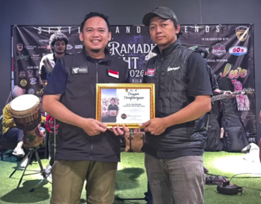 vespa60sjabodetabekdanscomadiindonesia7 dompet dhuafa Two men stand side by side on a stage, smiling, as they hold a framed award certificate together; a colorful event banner and musicians are visible in the background.