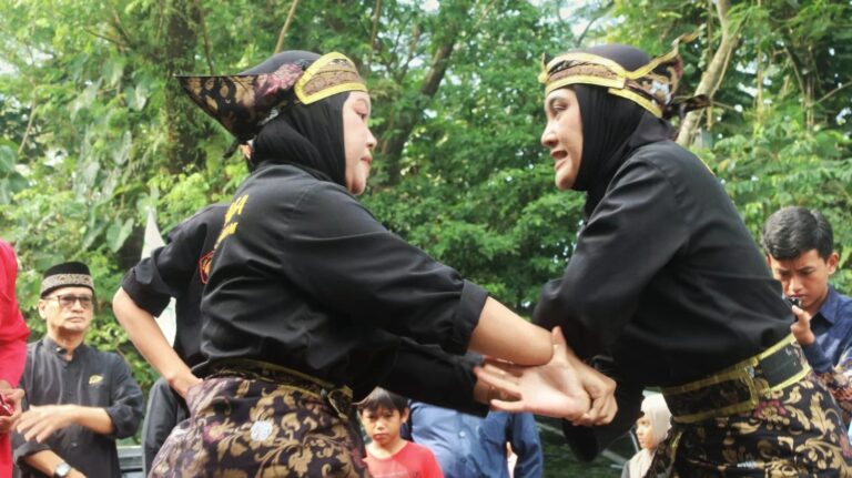whatsappimage20260427at141122768x431 dompet dhuafa Two women in black traditional outfits with gold sashes hold hands and dance outdoors as onlookers watch in a park-like setting.