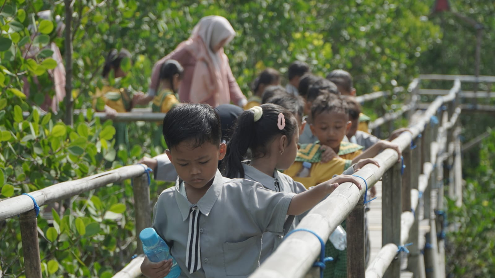 Group of children in uniforms walk along a bamboo suspension bridge amid dense green foliage, holding the railing.
