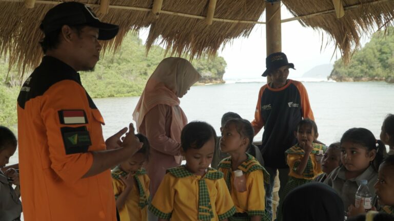 Group of Indonesian schoolchildren in yellow uniforms listening to a man in an orange uniform under a thatched roof near the water's edge.