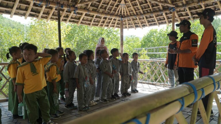 Group of schoolchildren in green and gray uniforms listening to two guides in orange shirts under a bamboo shelter.