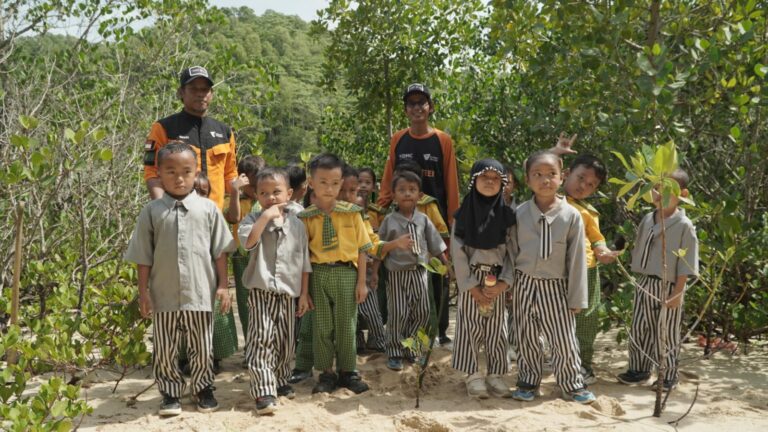 Group of children in gray shirts and striped pants with two adults in orange jackets posing on a sandy path among mangrove trees.
