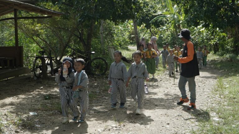 Children in striped pants walk single-file along a dirt path supervised by a guide in orange sleeves, outdoors among trees.