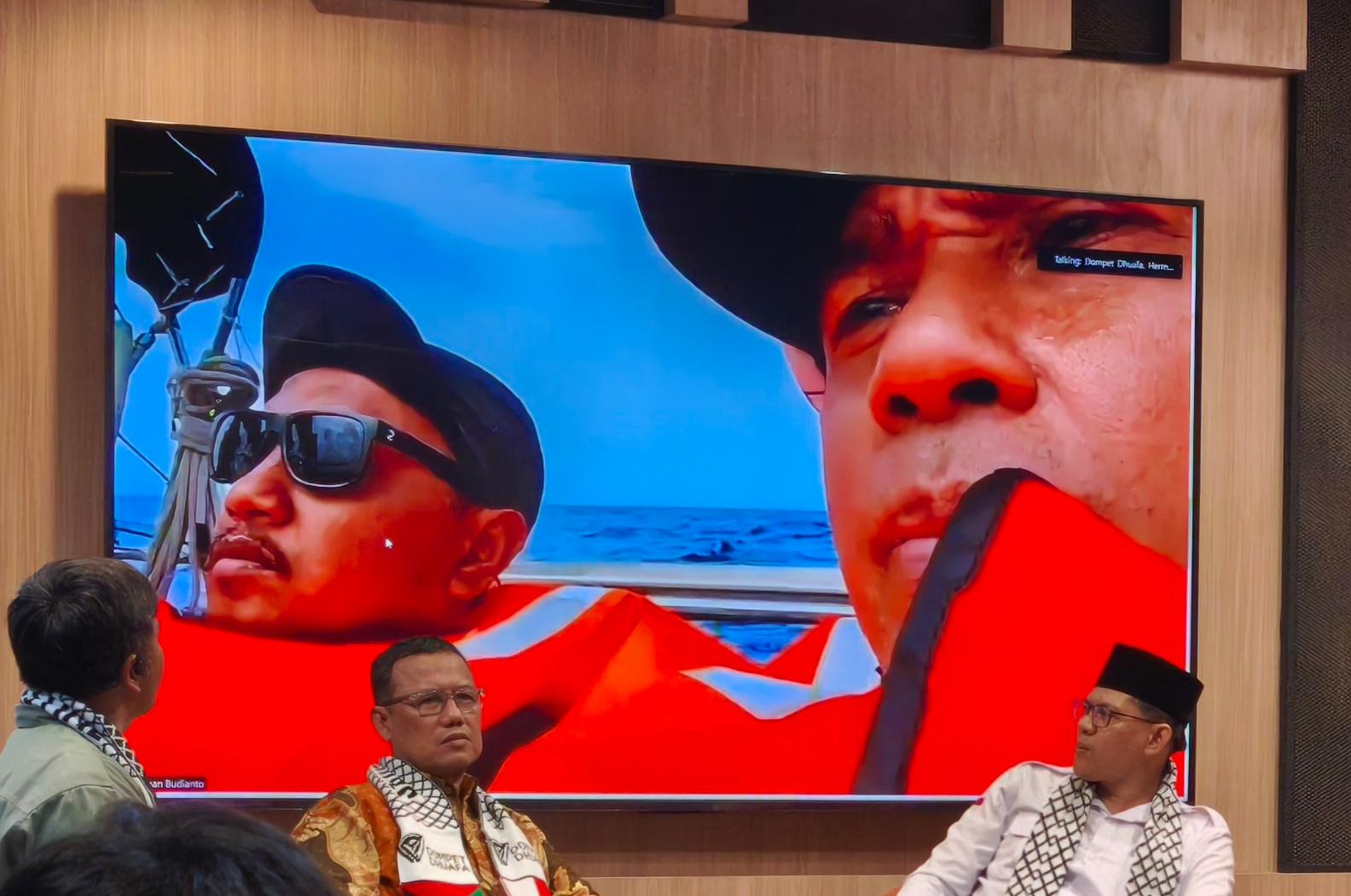 whatsapp image 20260520 at 181312 dompet dhuafa Large screen on a wooden wall showing close-up of two men in orange life jackets on a boat at sea; foreground, three men sit wearing scarves.
