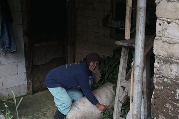 Woman in a blue jacket and teal pants crouches beside a large burlap sack outside a brick building, lifting or securing the sack.