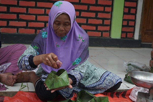 Elderly woman in a purple hijab sits on a red mat, folding a banana leaf to make a wrapped parcel in an outdoor market setting.