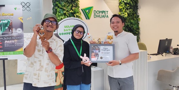 Three people pose in a Dompet Dhuafa reception area, smiling while holding plush toys and a framed certificate, with a green, plant-filled backdrop and logo in view.