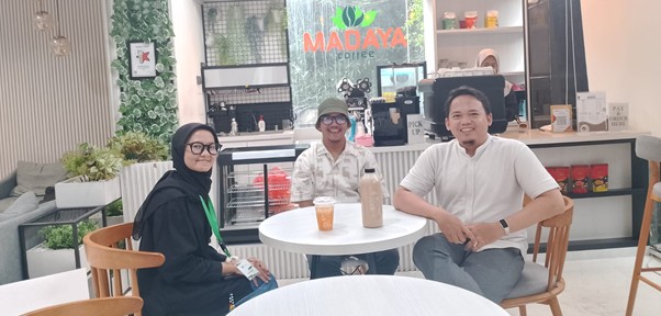 Three people sit around a white round table in a bright cafe, with beverages on the table and a MADAYA coffee sign visible in the background.