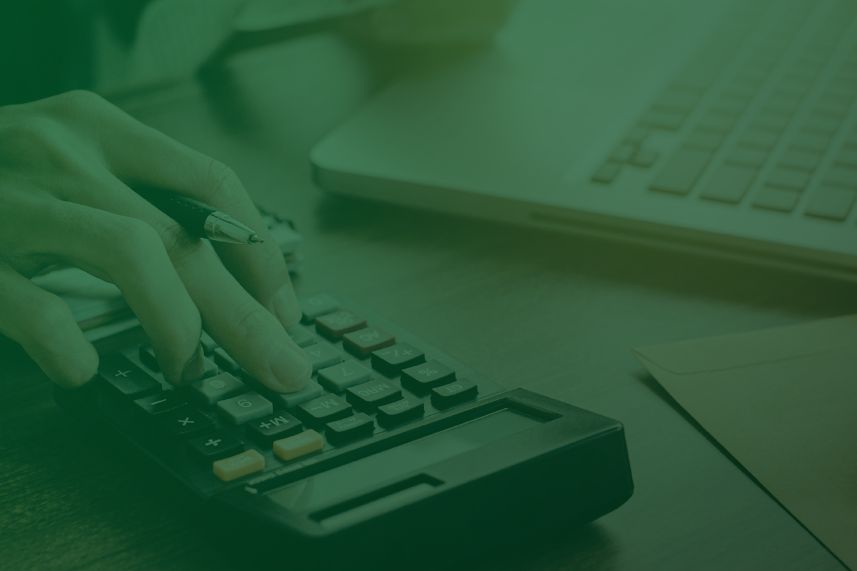 Person's hand pressing keys on a calculator with a laptop in the background on a desk, suggesting financial calculations.