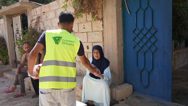 kurban di palestina 1 dompet dhuafa Volunteer in a hi-vis vest shakes hands with an elderly woman sitting by a stone wall outside a blue door.