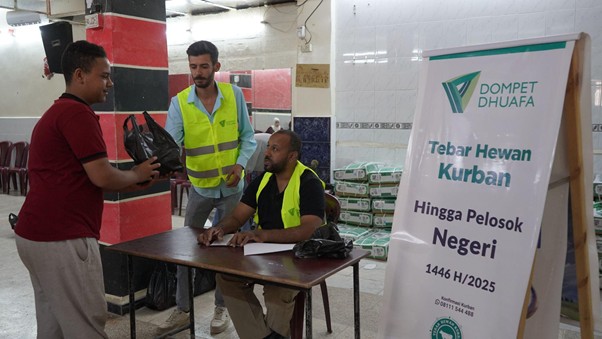 kurban di palestina 2 dompet dhuafa A man in a red shirt hands a bag to a volunteer seated at a table in a hall, with two volunteers in neon vests nearby and a Dompet Dhuafa banner in the background.
