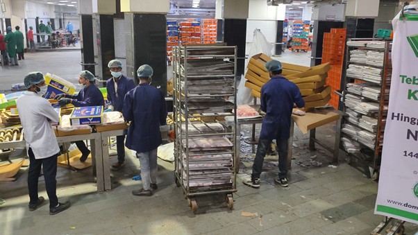 kurbandipalestina3 dompet dhuafa Factory workers in hairnets and blue uniforms pack pizza boxes in a busy food-processing area with carts and stacked boxes nearby.