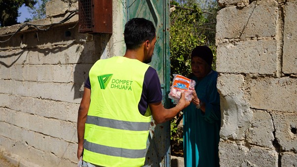 kurbandipalestina8 dompet dhuafa Aid worker in a high-visibility vest labeled DOMPET DHUAFA hands a package to an elderly woman at a doorway.