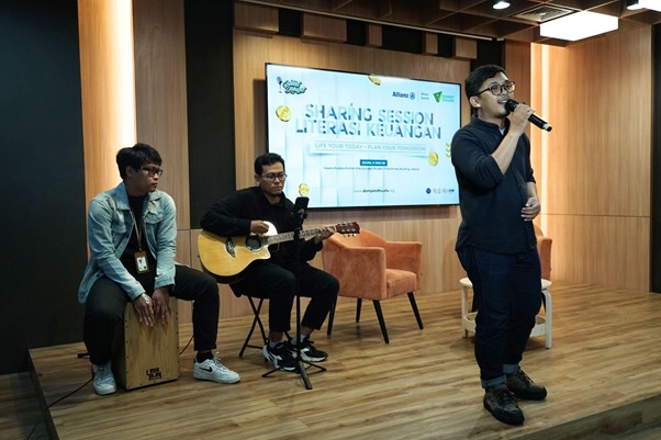 Three men perform on a wooden stage: a singer with a microphone standing, a guitarist, and a cajón player, with a screen that reads'Sharing Session Literasi Keuangan' in the background.
