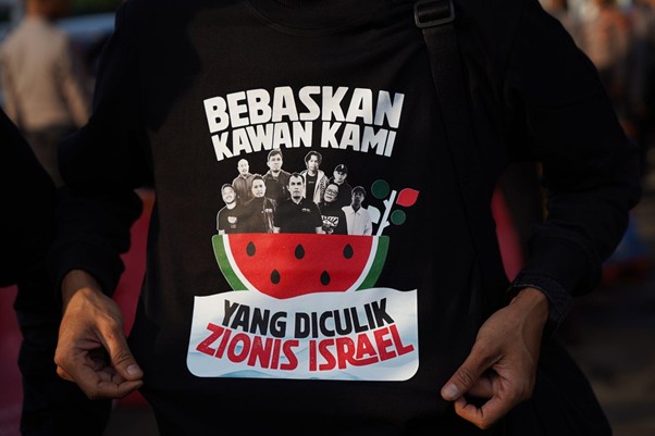 9wnibebasdariisrael2 dompet dhuafa Close-up of a black T-shirt with a protest graphic reading 'Bebaskan Kawan Kami' and 'Yang Diculik Zionis Israel' featuring a red watermelon and a crowd image.