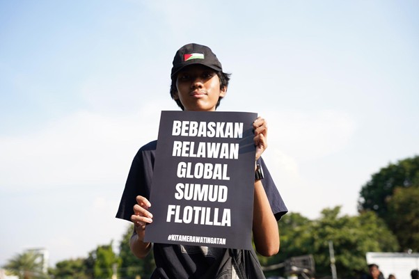 9wnibebasdariisrael3 dompet dhuafa Young person in a black cap holds a protest sign reading 'BEBASKAN RELAWAN GLOBAL SUMUD FLOTILLA' outdoors.