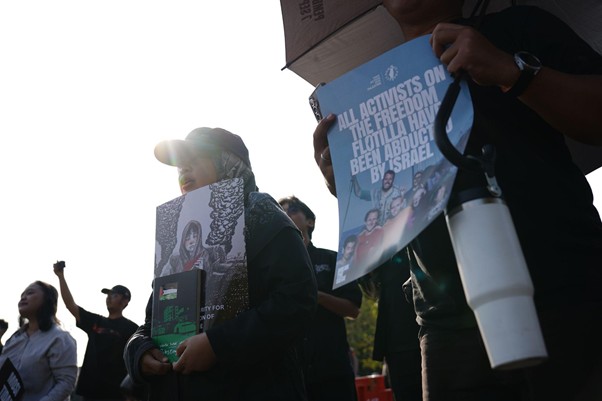 9wnibebasdariisrael4 dompet dhuafa Protesters at a demonstration holding signs; a blue poster with white text is visible in the foreground, sunlight behind them.