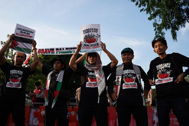 9wnibebasdariisrael5 dompet dhuafa Group of protesters holding signs at a rally, wearing black shirts and scarves, with a banner reading ‘Save Palestine’ in the background.