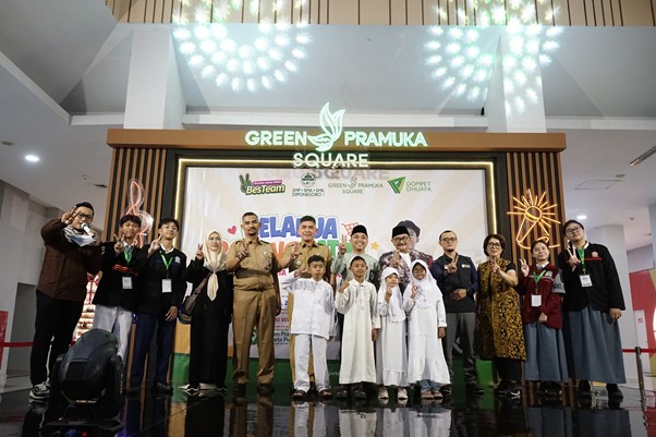 Group of children and adults posing on a stage at Green Pramuka Square during a community event, with banners and decorative lights in the background.