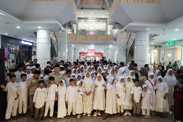 Large group of people, many children in white clothing, posing for a group photo in a shopping mall atrium.