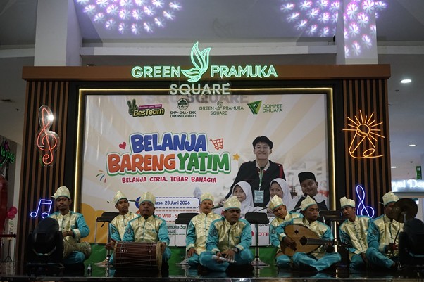 Group of men in turquoise outfits and traditional hats perform on a stage at a charity event, with a large Belanja Bareng Yatim banner in Green Pramuka Square mall.