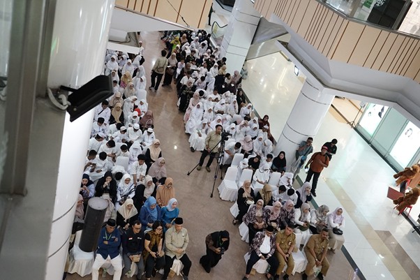 Large group of people seated in the atrium of a shopping mall, many wearing white robes, with a photographer on a tripod visible in the center.