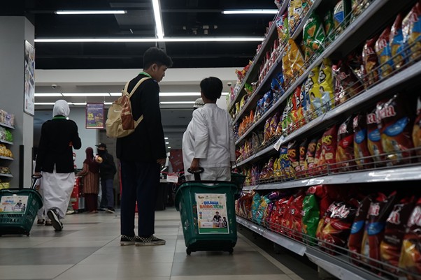 Two adults and a child with a green shopping cart browse snack shelves in a brightly lit supermarket aisle.