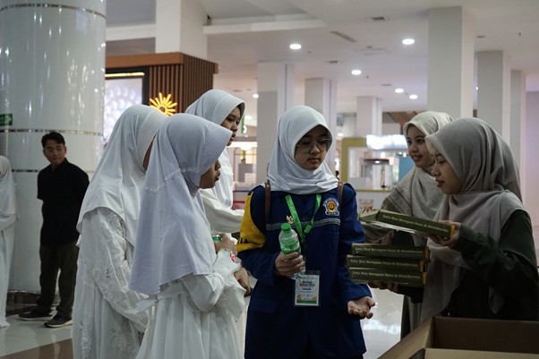 Group of women wearing hijabs in a bright indoor space discuss and share books, with a woman in a blue uniform holding a water bottle at the center.