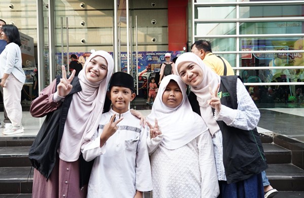 belanja bareng yatim prj 2026 3 dompet dhuafa Four people posing outside a glass-front building; two hijab-wearing women flank two children, all smiling and making peace signs.