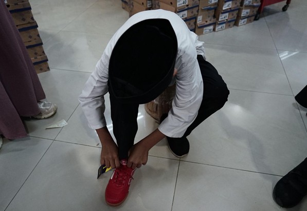 belanja bareng yatim prj 2026 4 dompet dhuafa Person kneeling on a light tile floor, tying the laces of a red sneaker, wearing a white top and black headscarf, with store shelves and boxes visible in the background.