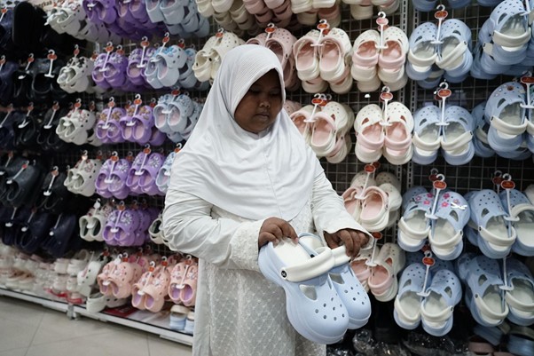 belanja bareng yatim prj 2026 6 dompet dhuafa Woman in a white hijab and dress holds a light blue pair of clogs while standing in front of a wall of colorful sandals.