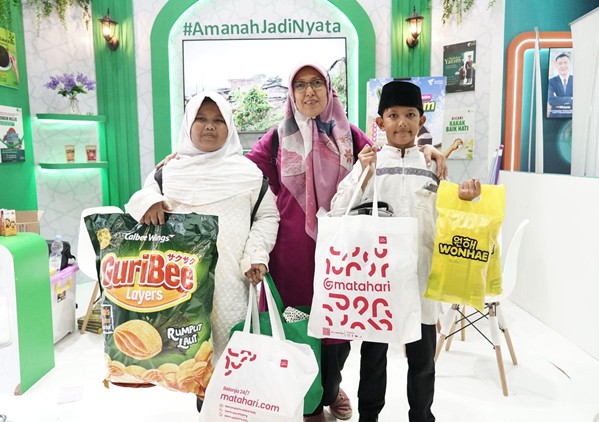 belanja bareng yatim prj 2026 8 dompet dhuafa Three people pose at a bright exhibition booth, each holding white or yellow shopping bags with logos visible; hashtag backdrop reads #AmanahJadiNyata.
