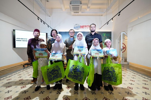 Group of children and adults posing with books and bright green bags at a donation event or charity program in a modern room with a screen displaying Indonesian text.