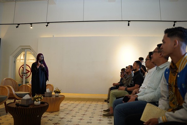 A woman in a hijab speaks to a seated audience in a bright, modern room with tiled floor and decorative tables and flowers in the foreground.