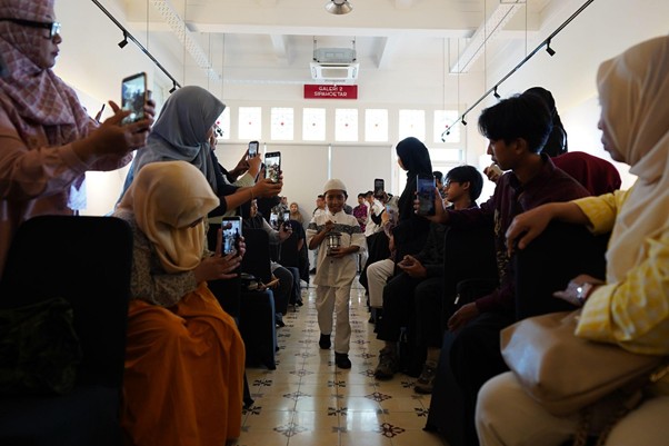 Young boy walking down an aisle between seated guests who are filming with their phones.