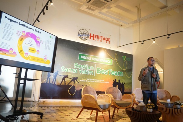 Speaker presenting at a press conference on a stage with a large banner reading 'HERITAGE CENTER' and a side monitor showing a colorful diagram.