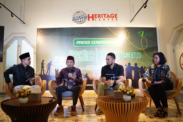 Panel discussion at a press conference on a stage with a banner reading 'Press Conference' and 'Heritage Center'; four people seated in chairs, two with microphones, two tables with flowers in front.