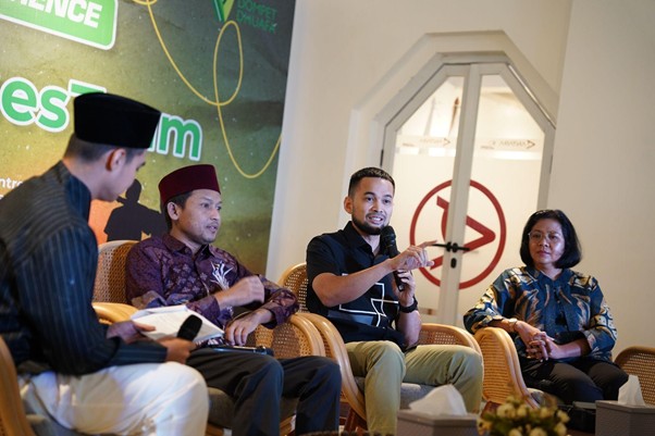 Panel discussion with four men seated in wicker chairs on stage; one man in a black shirt speaks into a microphone while others listen, with a green event banner in the background and a doorway with a red circular no-entry sign to the side.