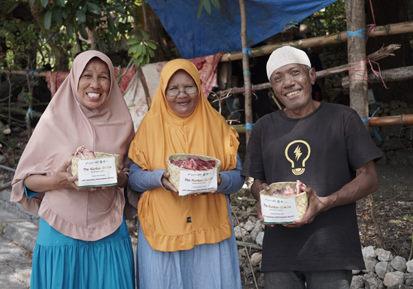 Two women in hijabs and a man in a white cap smile while holding donation boxes of meat outdoors.