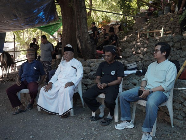 Four men sit on plastic chairs outdoors under a tarped shelter, with a stone wall and trees behind them.